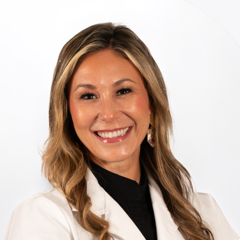 A woman with long blonde hair wearing a white coat and black top, smiling at the camera against a plain white background.