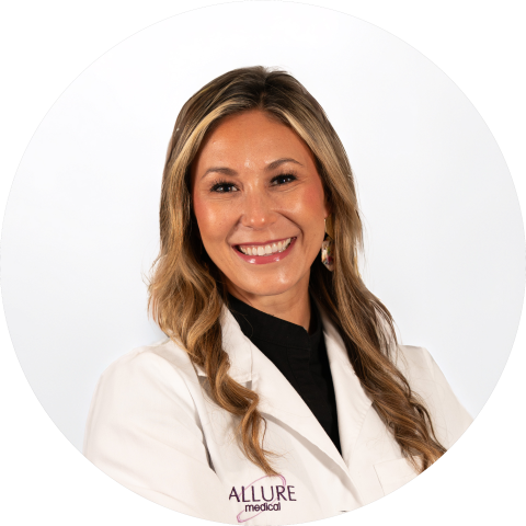 A woman with long, wavy hair smiles while wearing a white lab coat with "Allure Medical" embroidered on it, standing against a plain white background.