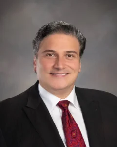 A man in a dark suit, white shirt, and red patterned tie smiles at the camera against a neutral grey background.