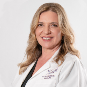 Woman with blonde hair wearing a white lab coat, smiling at the camera against a plain white background. The lab coat has "Allure Medspa" and a name embroidered on it.