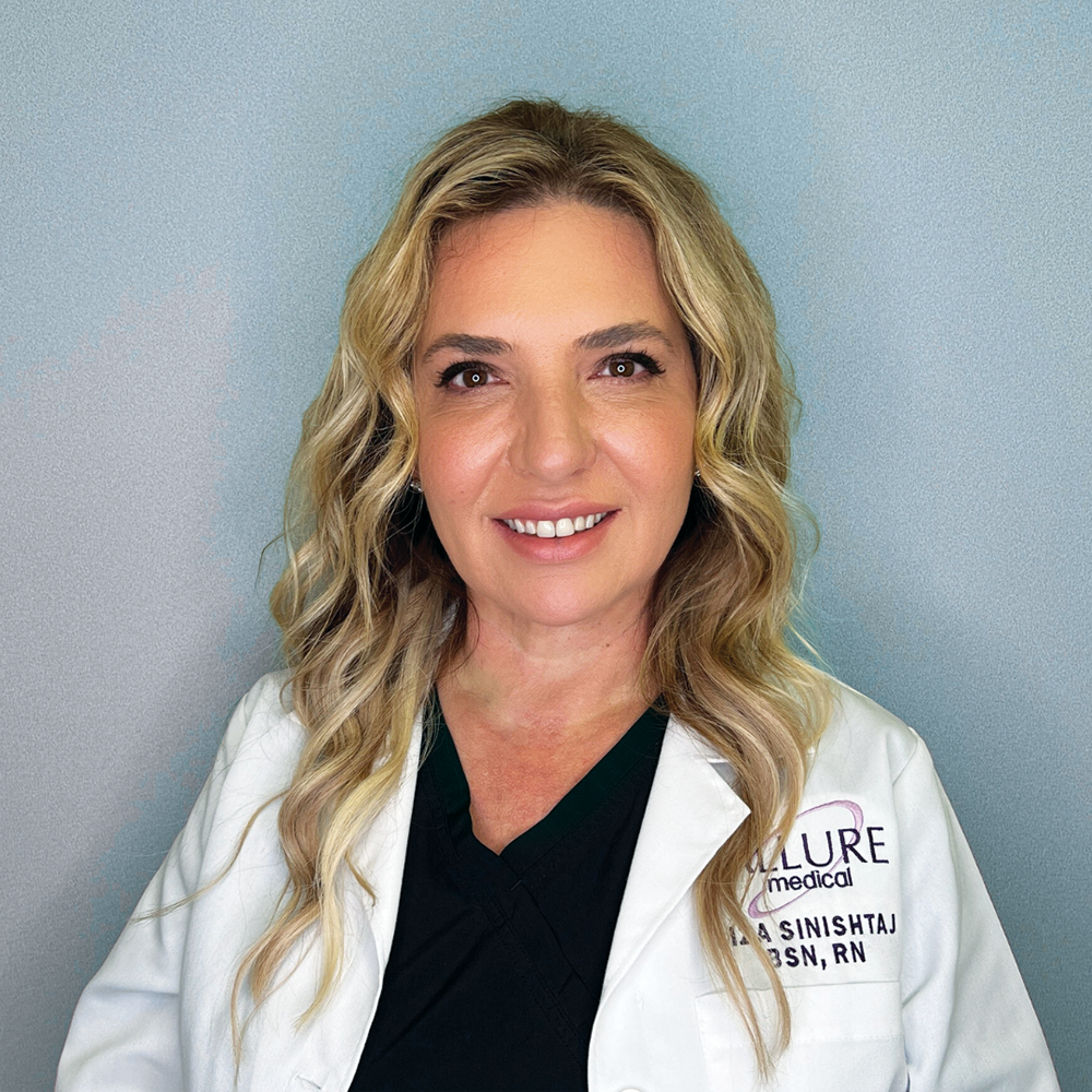 A woman with long blonde hair wearing a white medical coat over a dark top smiles at the camera. The background is a plain light blue.