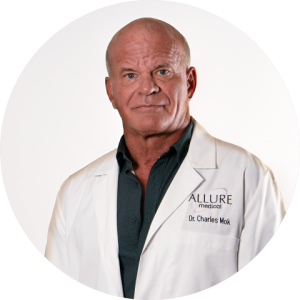 A man in a white lab coat with "ALLURE medical" and "Dr. Charles Mok" embroidered on it stands against a plain white background.