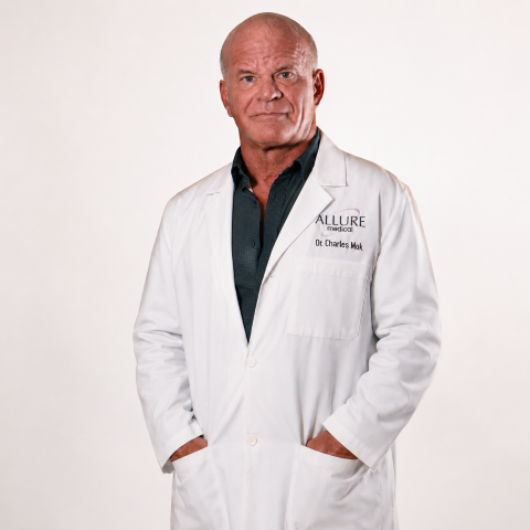 A man in a white lab coat with "Allure Medical" and "Dr. Charles Mok" embroidered on it stands with his hands in his pockets against a plain background.