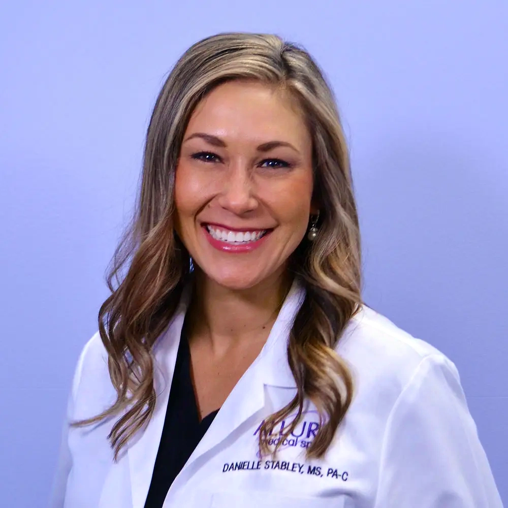 A person with long hair smiling, wearing a white lab coat with text reading "DANIELLE STABLEY, MS, PA-C" and "ALLURE Medical Spa" against a plain background.