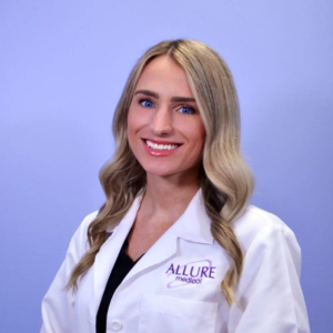 A woman with long blonde hair and blue eyes, wearing a white lab coat with "Allure Medical" embroidered on it, smiles against a light blue background.