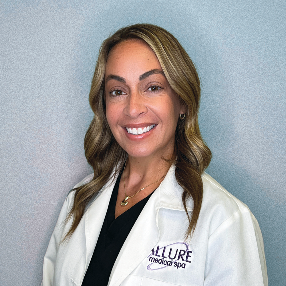 A person with long, wavy hair wearing a white coat and a black shirt stands against a neutral background, smiling. The coat has "Allure Medical Spa" embroidered in purple.