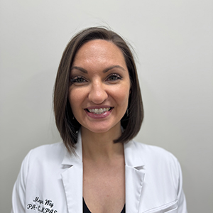 A person with shoulder-length brown hair smiles at the camera, wearing a white lab coat and a black top, standing against a plain white background.