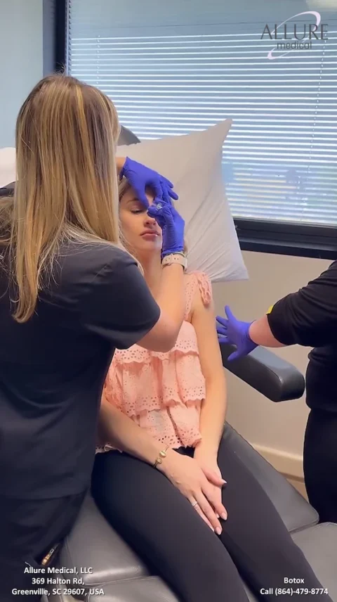 A woman receives a Botox injection on her forehead at a medical clinic. Staff members wearing gloves administer the treatment.