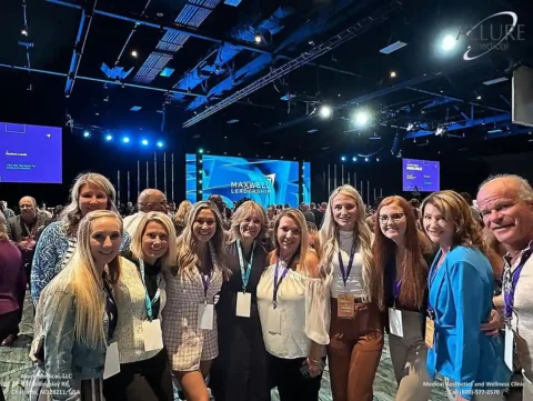 Group of people posing together and smiling in a large indoor event space with a stage in the background displaying the text "MAXWELL LEADERSHIP.