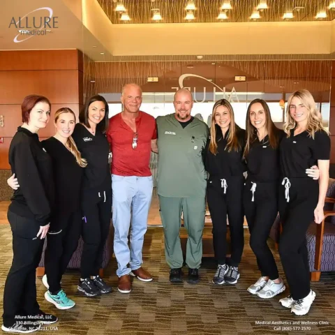 A group of seven people, including six women in medical attire and two men, one in red shirt and the other in green scrubs, standing together in a medical clinic lobby with Allure Medical sign.