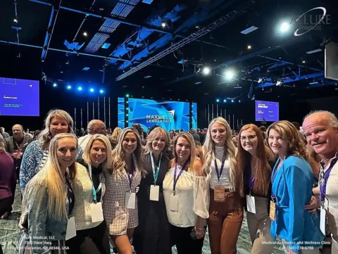 A group of people smiling for a photo in front of a stage at what appears to be a conference or event. They are indoors with bright overhead lights and several display screens in the background.