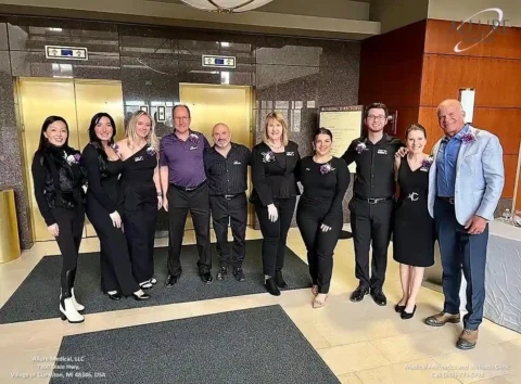 A group of nine people, dressed in business attire, standing in a lobby area with elevators in the background. Some are wearing black uniforms, and others are in formal wear. An informational sign is visible.