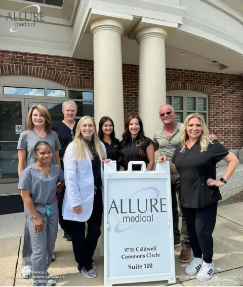 A group of eight people, all dressed in various medical uniforms, stand smiling outside a medical building next to a sign for Allure Medical at 9735 Caldwell Commons Circle, Suite 100.