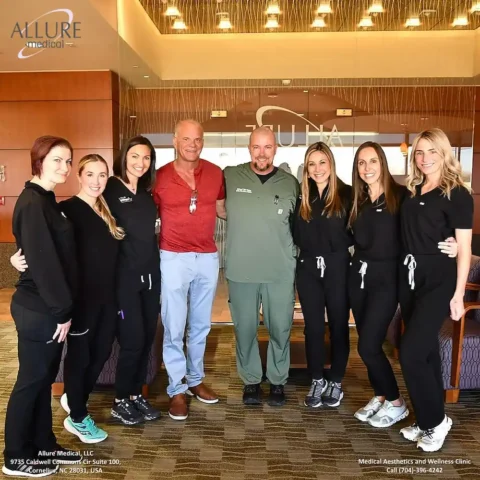 A group photo shows seven people standing inside a clinic. There are two men in the center, one in a red shirt and another in medical scrubs, surrounded by five women in matching black uniforms.