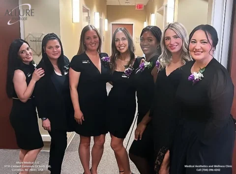 A group of seven women dressed in black stand together in a hallway, smiling at the camera. The wall behind them displays the Allure Medical logo and contact information.