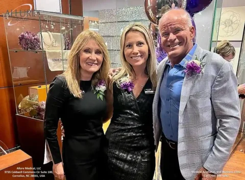 Three people stand closely together, smiling. They are dressed formally and each wears a floral boutonniere. A sign in the background reads "Allure Medical.