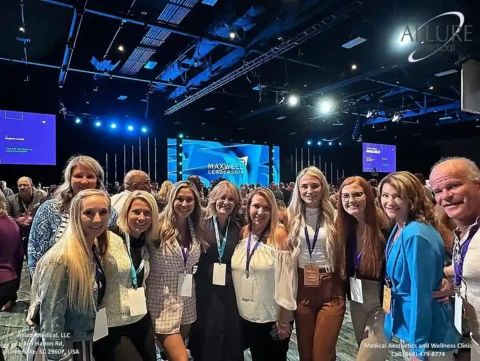 A group of people with conference badges pose for a photo in a large, well-lit auditorium with multiple display screens and stage lighting. The event appears to be a professional conference.