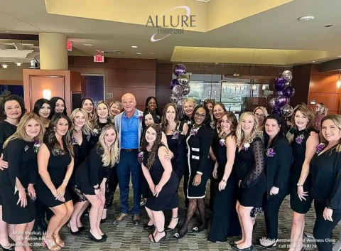 A group of people, predominantly women dressed in black, stand together in an indoor setting. A man in a blue shirt and vest is at the center. Purple balloons decorate the background.