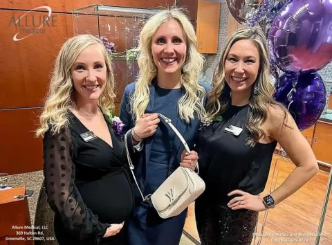 Three women smiling and posing together indoors. The center woman holds a white purse with a "LV" logo. Purple balloons are visible in the background.