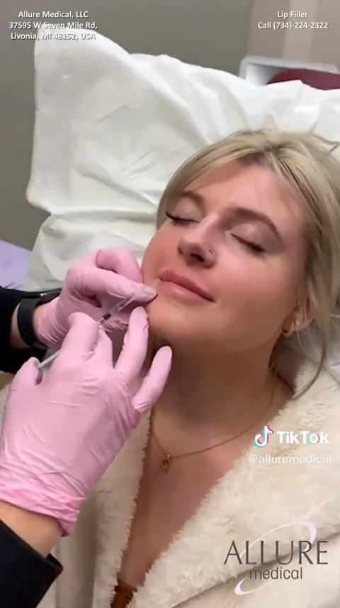 A woman receives a lip filler injection from a person wearing pink gloves in a clinical setting.