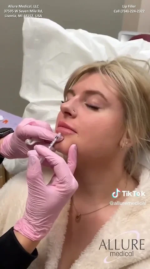 A woman receives lip filler injections at Allure Medical clinic. A practitioner with pink gloves is administering the injection while the woman lies back with her eyes closed.