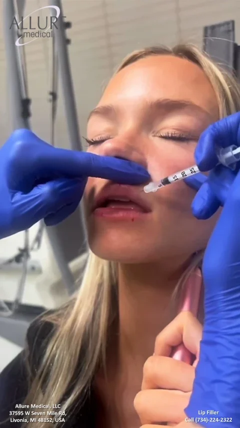 A person receives a lip filler injection from a gloved medical professional. The clinic's name, address, and contact information are shown at the bottom of the image.