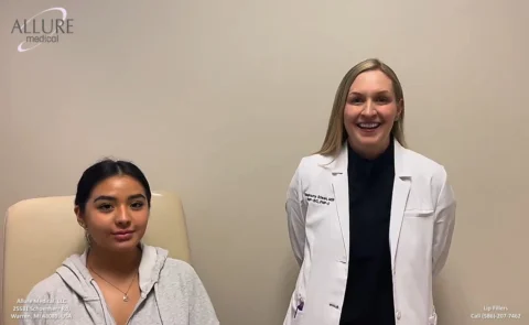 A medical professional in a white coat stands next to a seated patient in a clinic exam room. The patient is wearing a light gray hoodie. The background wall is plain and light-colored.