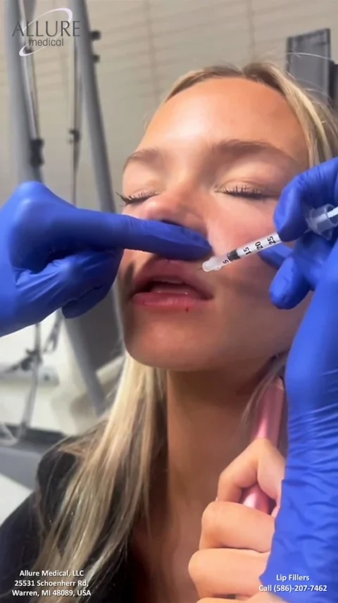 Person receiving lip filler injections from a medical professional. Blue-gloved hands are holding a syringe to the person's lips. Signage in the background shows the clinic's name and contact information.