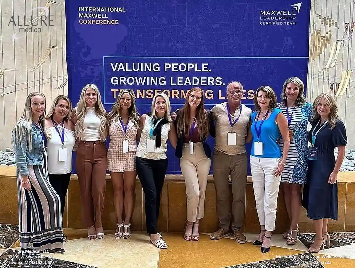 A group of people stands in front of a backdrop at the International Maxwell Conference. The backdrop reads, "Valuing People. Growing Leaders. Transforming Lives." They are posing for a photo.