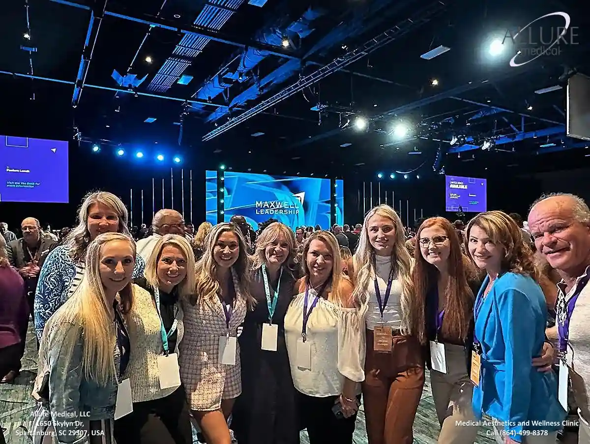 A group of people posing for a photo at a conference venue with stage and screens visible in the background. The room is dimly lit with blue-toned lighting.