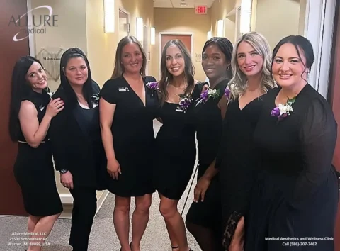 Six smiling women in black outfits stand in a hallway, displaying name tags from "Allure Medical." Some wear white and purple flower corsages. Walls feature warm tones and the clinic’s name and details are visible.