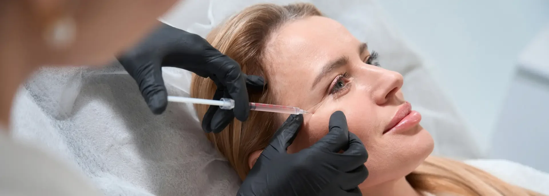 A woman receives a facial injection from a professional wearing black gloves in a medical setting.
