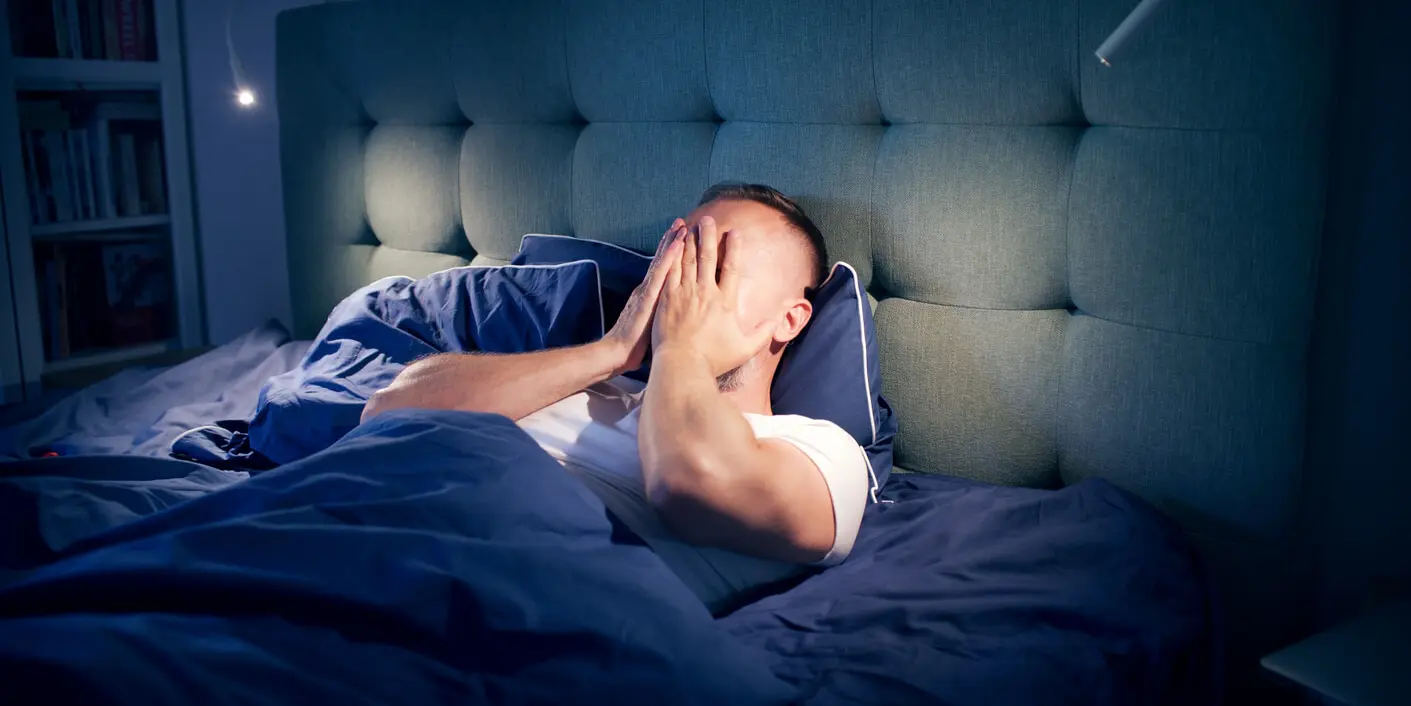 A person sitting in bed with a blue headboard, covering their face with both hands while dressed in a white shirt and blue bedding.
