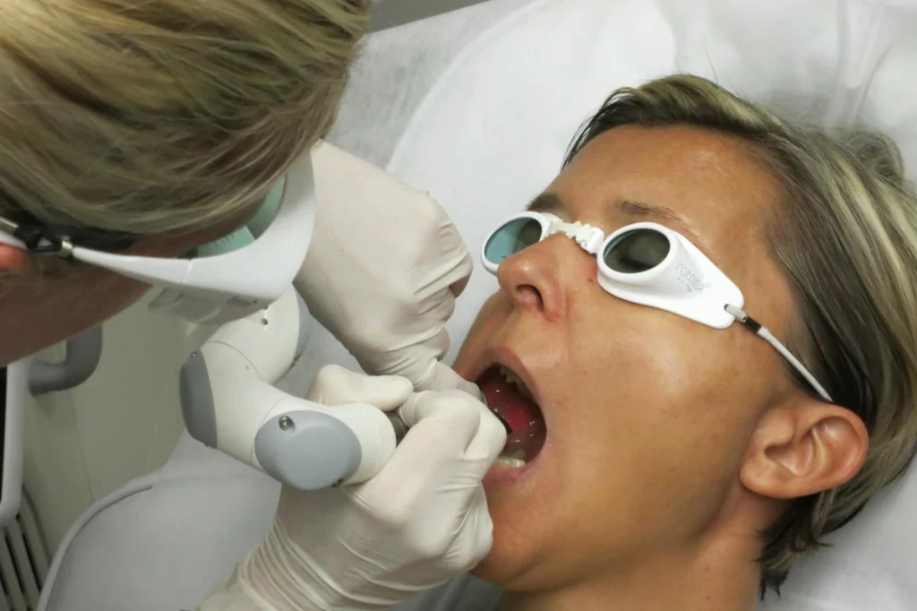 Person receiving dental treatment with protective goggles, while a dentist uses a laser tool in a clinical setting.