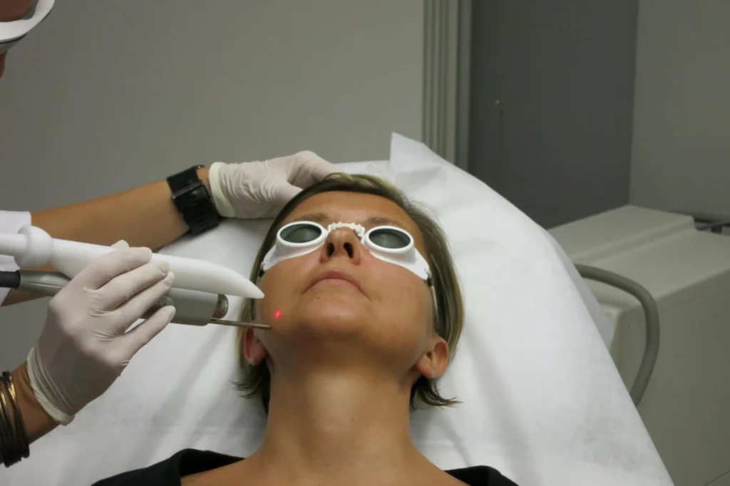 A person wearing protective goggles receives a laser treatment on their chin from a gloved professional.