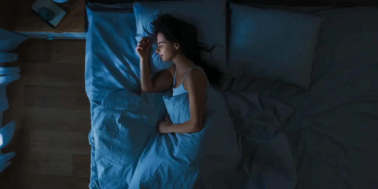 A woman sleeps on a bed at night, covered with a light blue blanket. The room is dimly lit with a nightstand visible on the left side of the image.