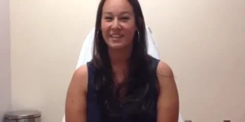 A woman with long dark hair sits smiling in a chair against a beige wall. She is wearing a dark sleeveless top and has hoop earrings.