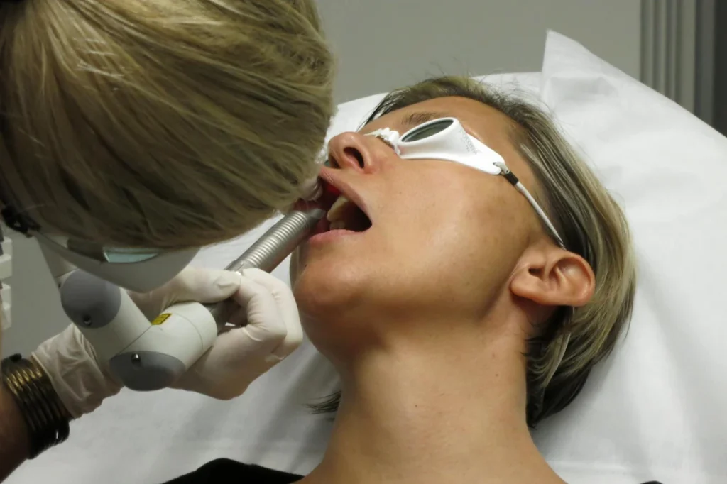 A person receives a dental treatment with a handheld device while wearing protective goggles in a clinical setting.