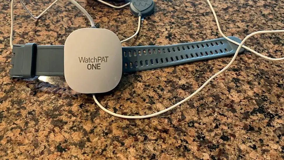 A WatchPAT ONE device with a wrist strap and connected sensors is placed on a granite countertop.