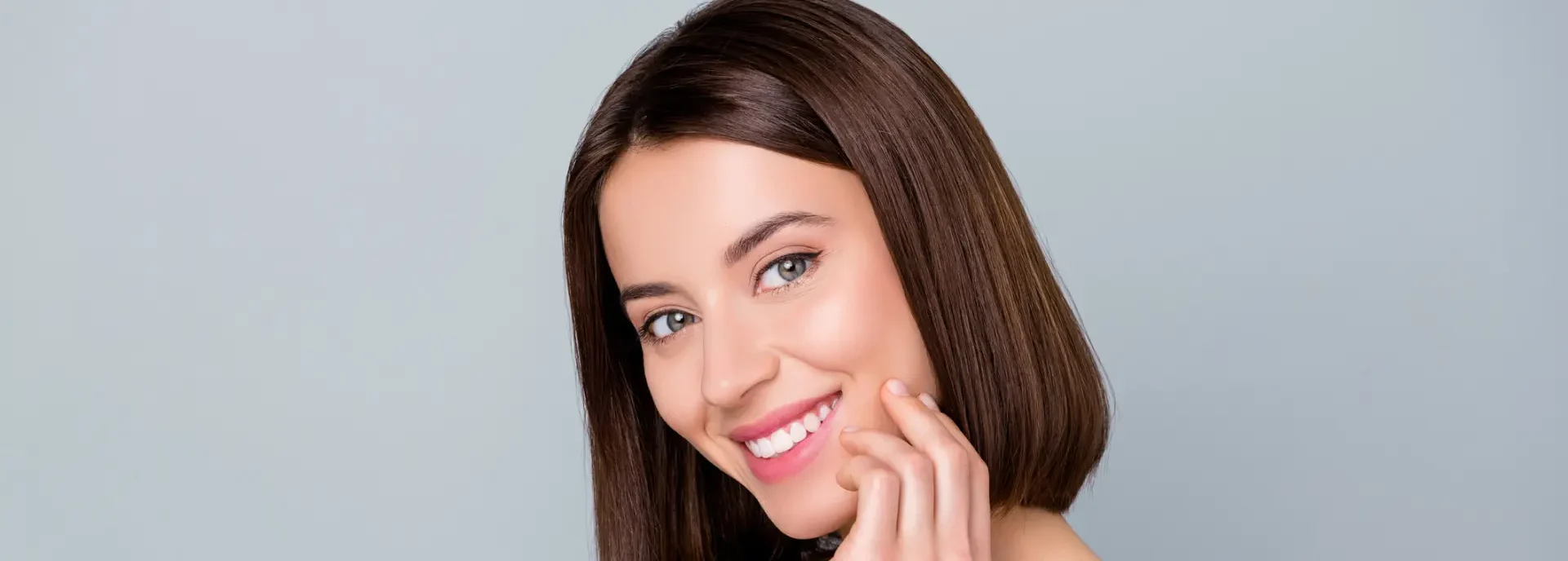 A woman with straight brown hair and fair skin is smiling, with her hand gently touching her chin, against a plain light gray background.