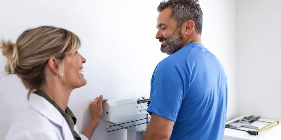 A man in a blue shirt stands on a medical scale while a woman in a white coat smiles and observes in a clinical setting.