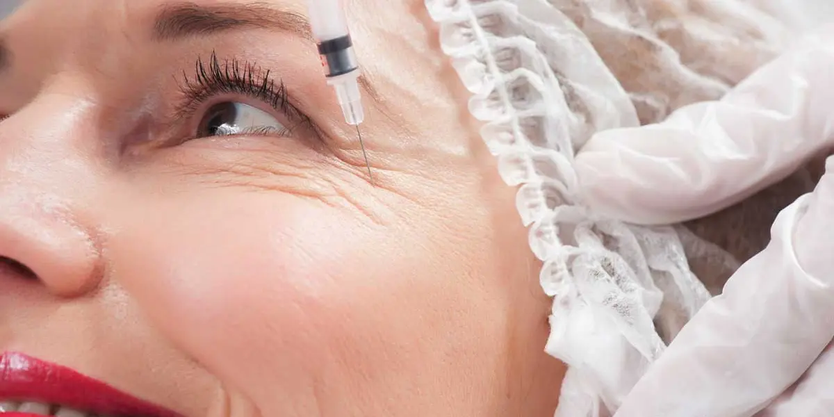 A close-up of a woman's face receiving a cosmetic injection near her eye. She is wearing a hair net and gloves are visible on the left side of the image.