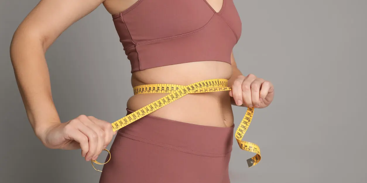 Person in sportswear measuring their waist with a yellow measuring tape against a gray background.
