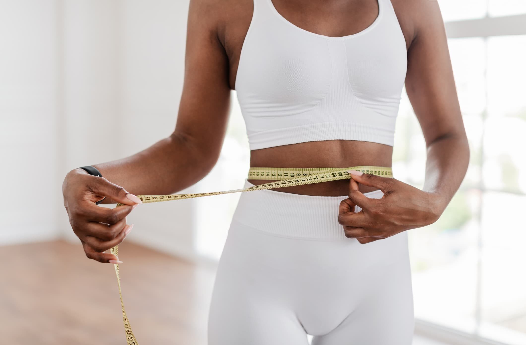 A person in a white fitness outfit measures their waist with a yellow measuring tape in a bright, indoor setting.