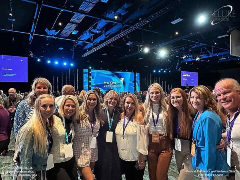 Group of people standing together and smiling at a conference event with a large screen in the background displaying "Maxwell Leadership" text.