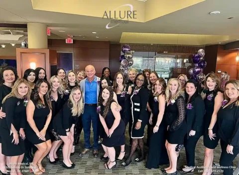 A group photo of twenty-three people, including a man in a blue suit and women in black dresses, posing inside a modern office setting with purple balloons and the Allure Medical logo.