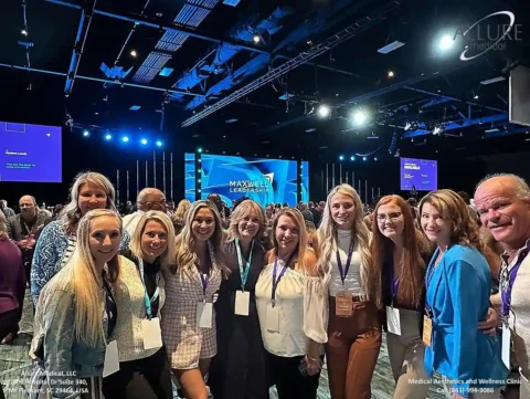 A group of people pose for a photo at the Maxwell Leadership conference inside a large convention hall. A stage with a screen displaying "Maxwell Leadership" is visible in the background.