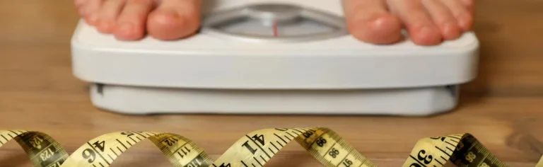 Bare feet standing on a bathroom scale with a yellow measuring tape coiled on the floor in front.