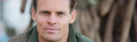 A man with short hair and blue eyes looks directly at the camera, wearing a green jacket. The background is blurred.