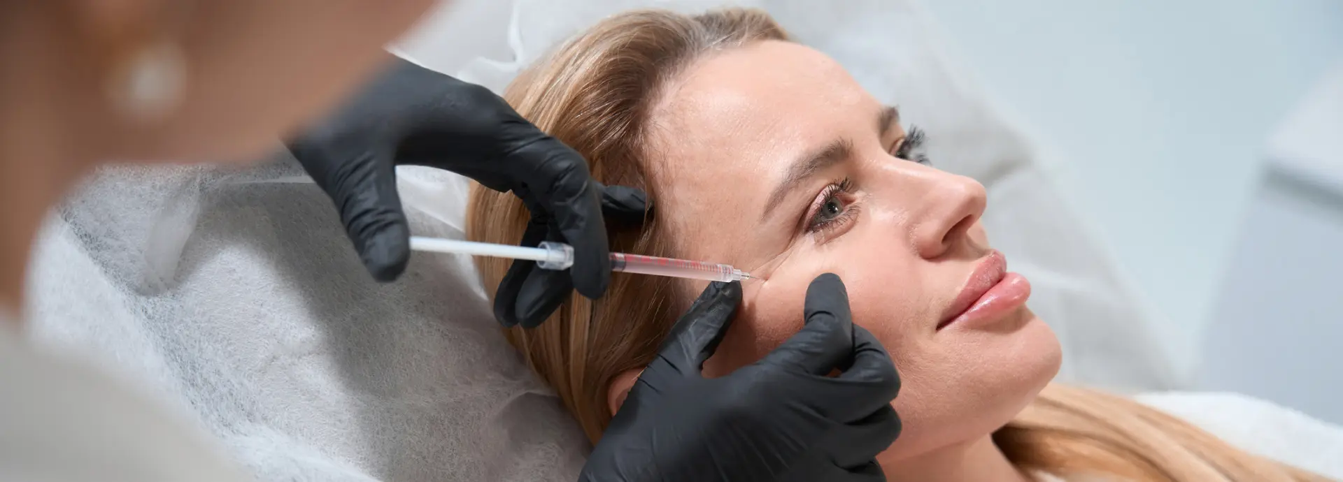 A person with black gloves administers an injection to a woman's face as she reclines on a medical chair.